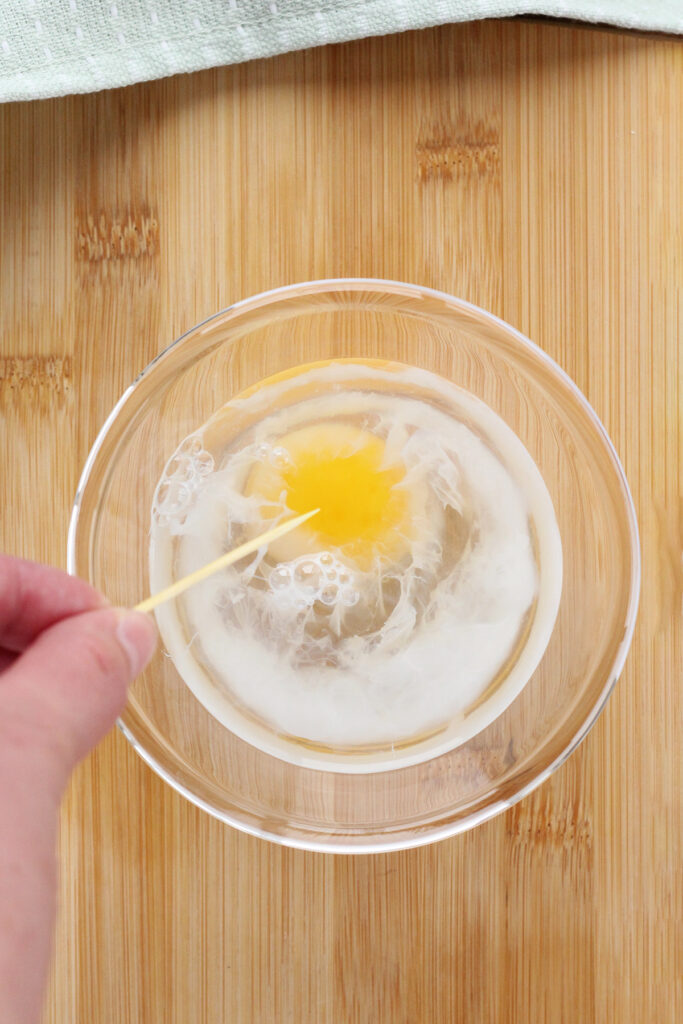 an egg inside a glass dish of hot water with the yolk being pricked by a toothpick