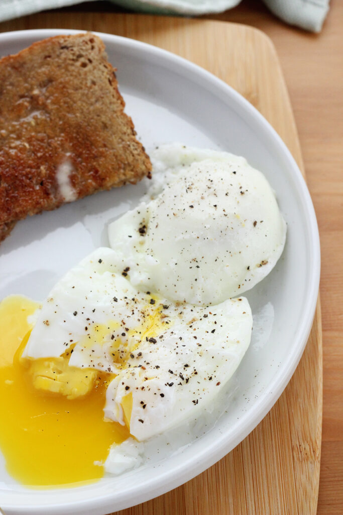 close up image showing two poached eggs seasoned with salt and pepper on a small white round plate with toast off in the background