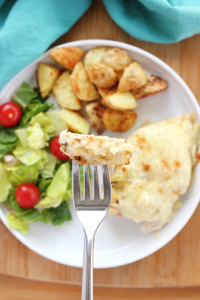 close up image showing a chicken breast topped with artichoke dip and cheese with a bite on a fork. The chicken is on a white round plate with a side salad and crispy potatoes