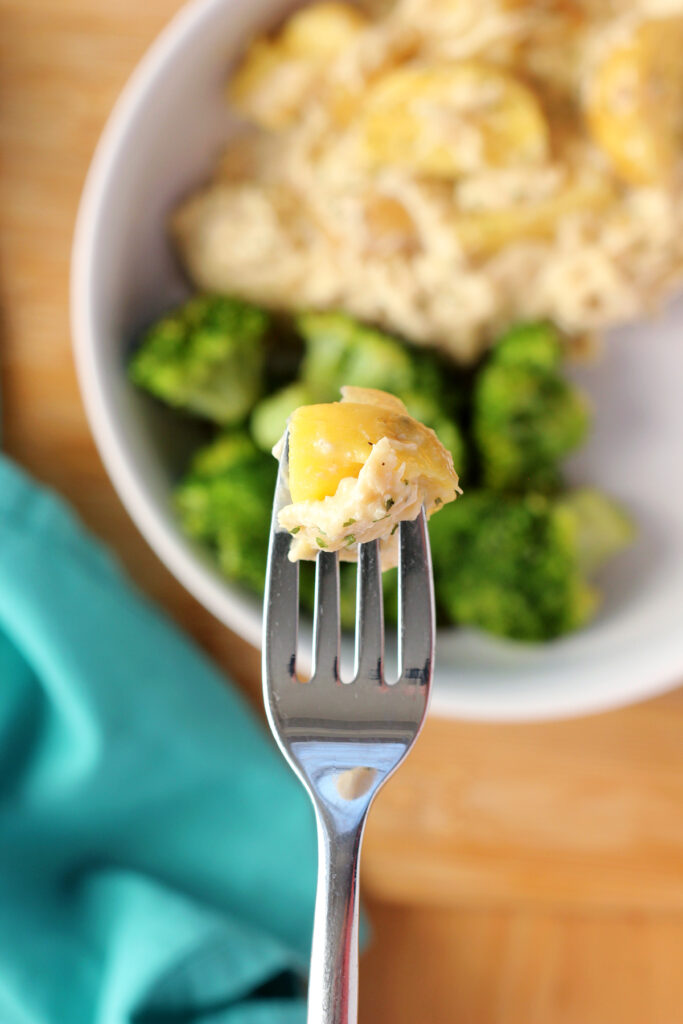 close up image showing a shallow bowl filled with shredded chicken and potatoes in a sauce with roasted broccoli. A bite is being held over the plate on a fork