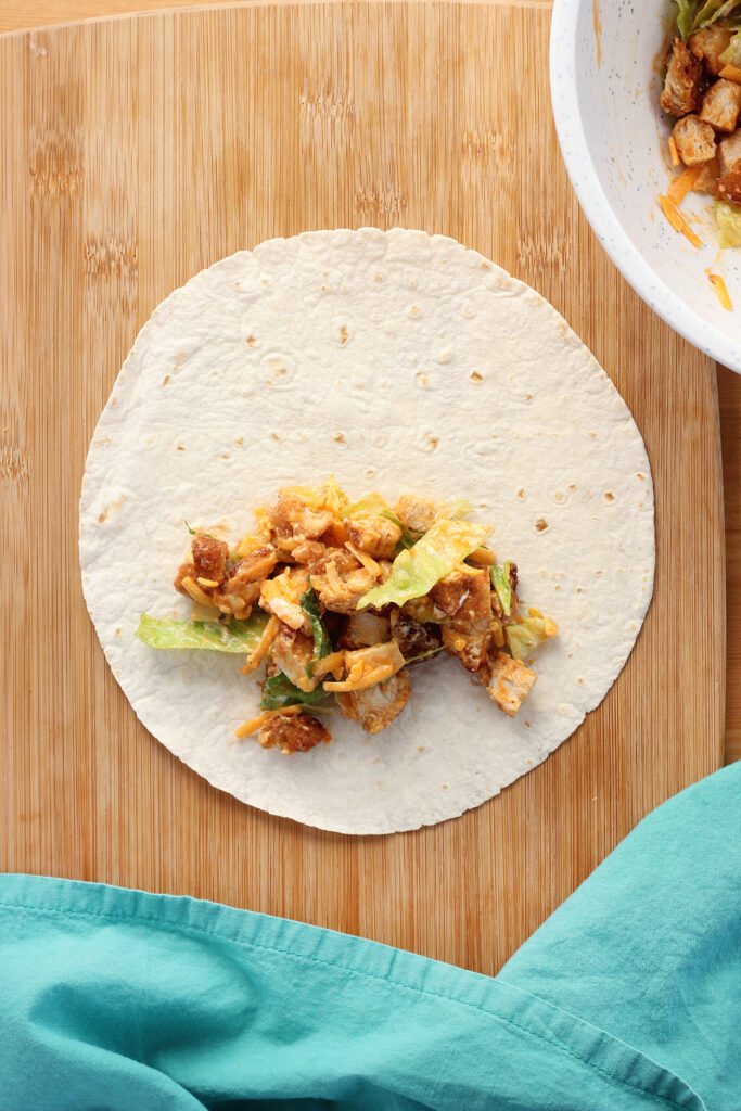 top down image showing a tortilla on a cutting board with chicken wrap filling sitting on top