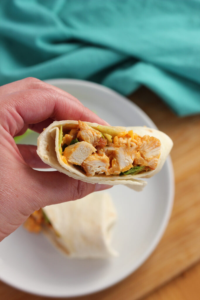 hand holding half of a chicken wrap over a plate with the other half. A teal napkin is off in the back. 