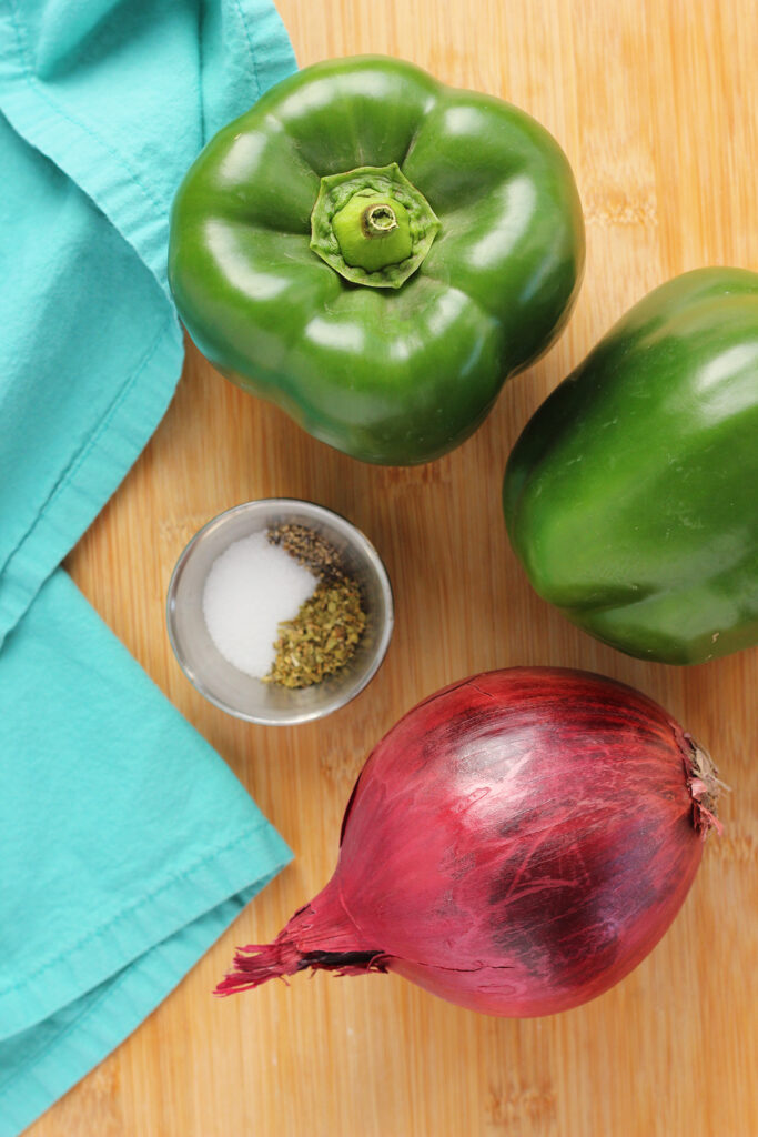 top down image showing two green bell peppers, one red onion, and a small container with salt, pepper, and oregano sitting on a wooden cutting board with a teal napkin