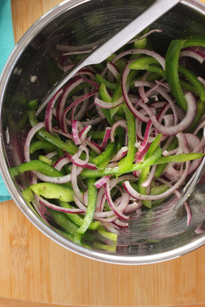 top down close up image of a metal bowl filled with mixed green bell pepper and red onion being tossed with oil and seasoning by metal tongs