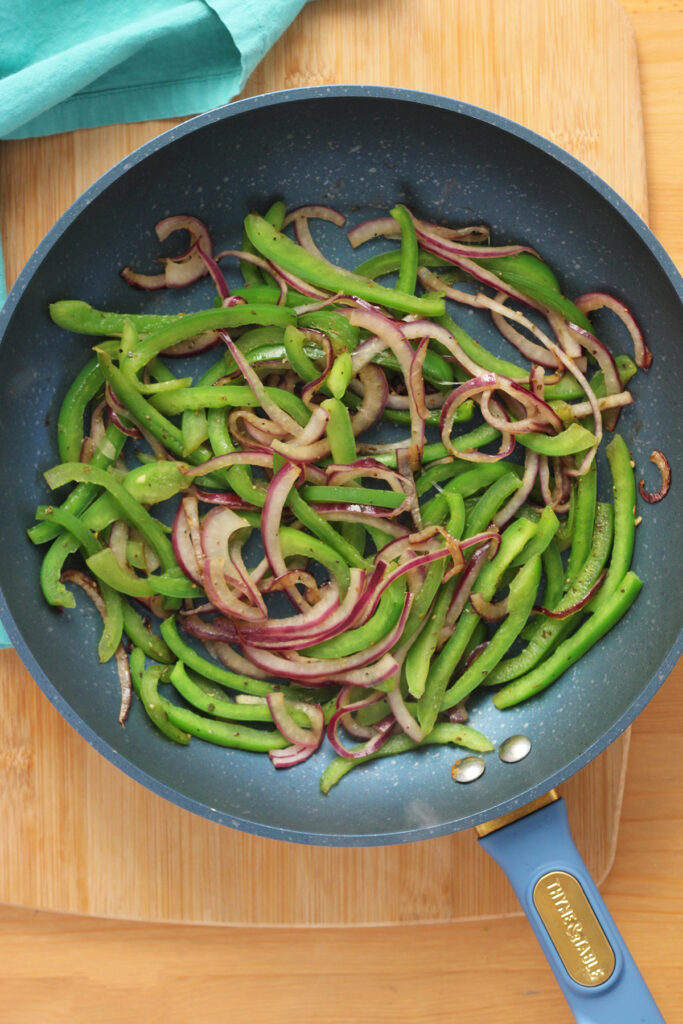 upclose image of a blue frying pan showing cooked green bell pepper and red onion slices. The pan is sitting on a wooden cutting board with a teal napkin