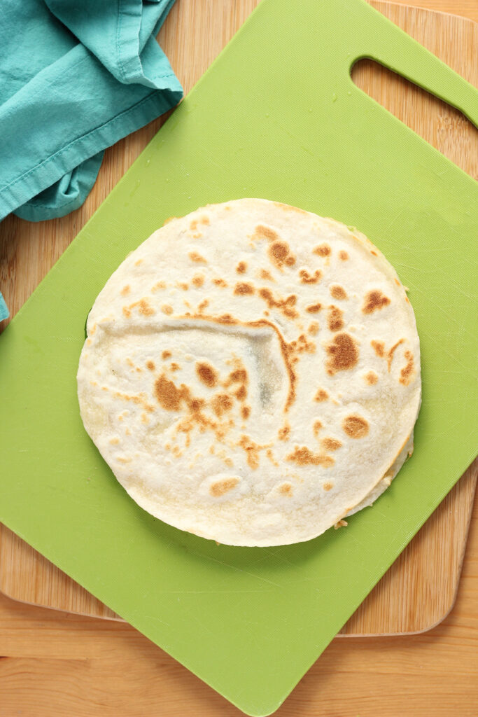 top down image showing a cooked quesadilla sitting on a green cutting board ready to be cut. 