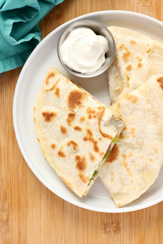 close up image of quesadilla slices stacked on each other on a white round plate with a small dish of sour cream off in the background with a teal napkin