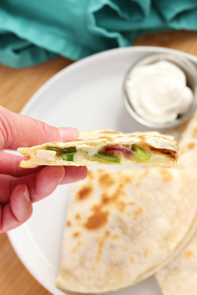 hand holding a quesadilla triangle over a white round plate that is holding more pieces of the quesadilla with a side of sour cream.