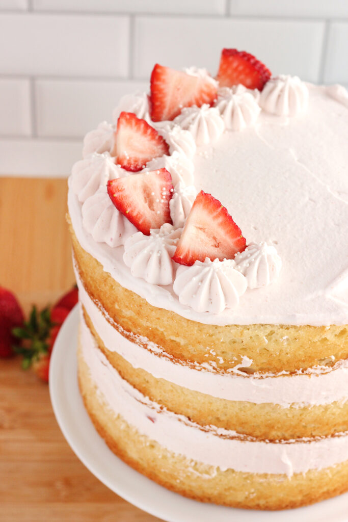 angled top down image showing a three layer white cake that is filled with pink whipped cream and topped with strawberry slices. The cake is sitting on a white cake stand with fresh strawberries scattered around on a wooden table