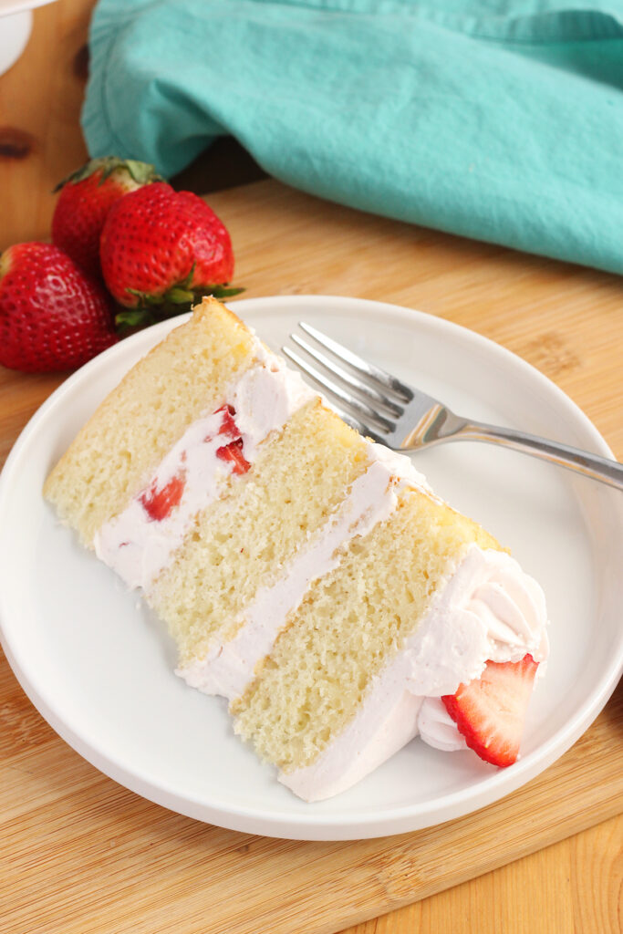 close up image of a slice of white caked layered with pink whipped cream sitting on a white round plate with a fork. The plate is sitting on a wooden table top with fresh berries behind it