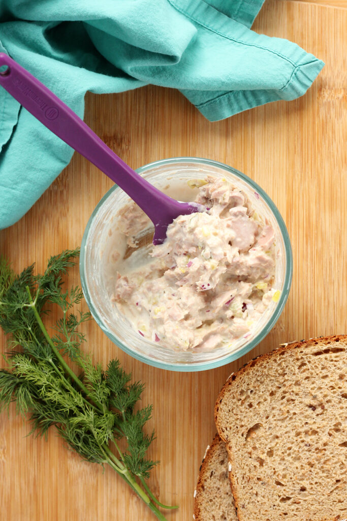 Top down image of a glass bowl filled with tuna salad and a purple silicone spatula. The bowl is sitting on a wooden cutting board with two slices of bread, fresh dill, and a teal napkin