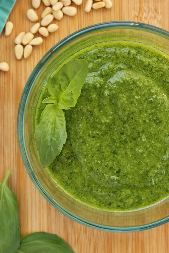 close up image of a glass bowl filled with pesto with fresh basil leaves sitting on top. The bowl is sitting on a wooden table top with more basil, pine leaves, and a teal napkin off to the side