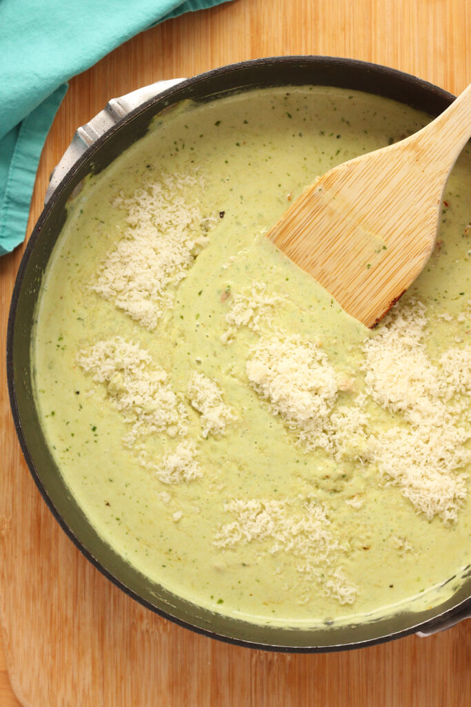 Top down image showing a black skillet filled with a green sauce and sprinkled with parmesan cheese. There is a wooden spatula inside the pan 