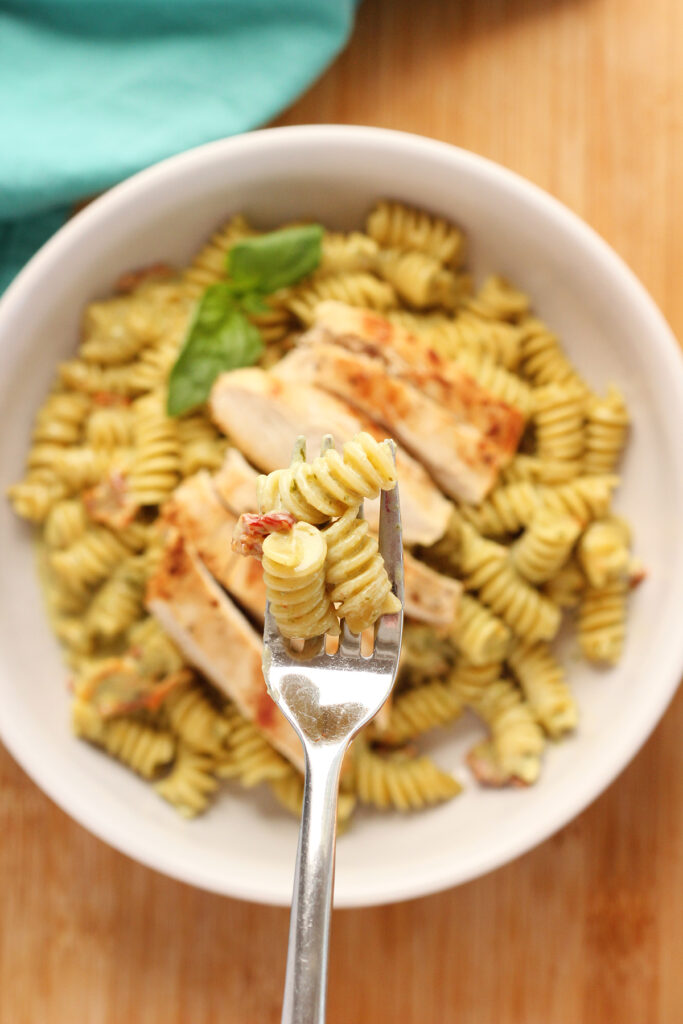 close up top down image showing a shallow white bowl filled with rotini pasta that is covered in a light green sauce with sun dried tomatoes sprinkled throughout. A fork is holding a bite over the top
