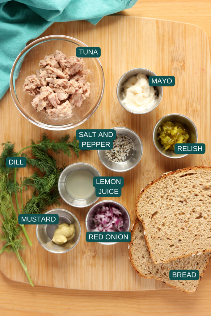 Top down image of a wooden cutting board showing a bowl of tuna, small dish of mayo, small dish of relish, salt and pepper, small dish of lemon juice, small dish of mustard, small dish of finely diced red onion, two sliced of bread, and fresh dill with a teal napkin
