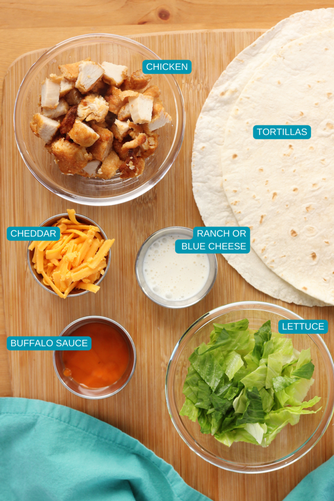 top down image showing a wooden cutting board topped with a glass bowl of crispy chicken chopped, tortillas, bowl of cheddar, bowl of ranch, bowl of buffalo sauce, and a bowl of lettuce