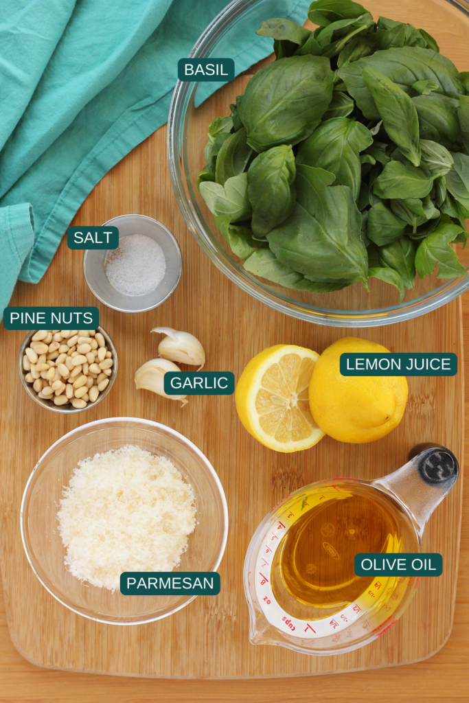 top down image showing a glass bowl of basil, small dish of salt, small dish of pine nuts, two cloves of garlic, a lemon sliced in half, small dish of parmesan cheese, and measuring cup of olive oil sitting on a wooden table top with a teal napkin. All ingredients are labeled with a text overlay