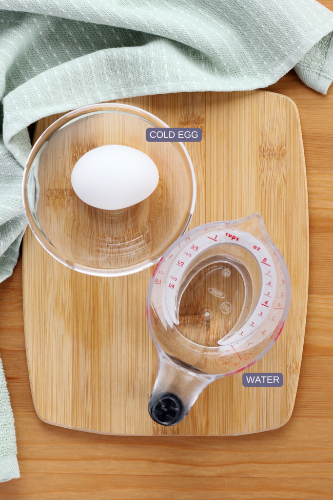 top down image showing a wooden cutting board with a small glass bowl with a single egg inside and a measuring cup filled wiht water. There is a light blue napkin off to the side