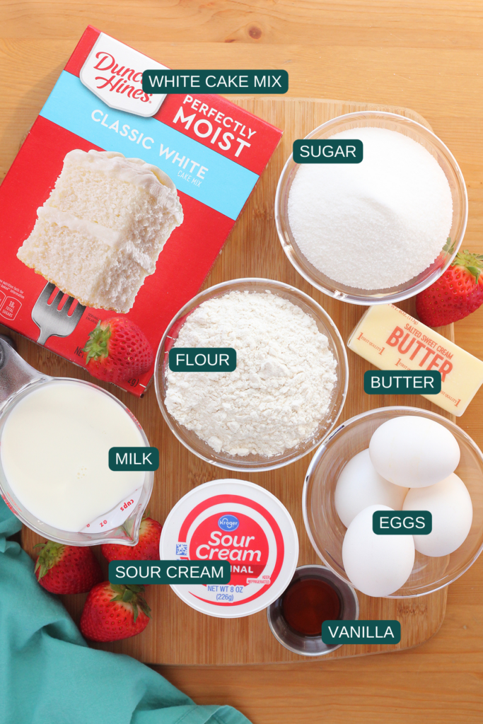top down image showing a wooden table top that has a box of duncan hines classic white cake mix, a bowl of sugar, a bowl of flour, a stick of butter, bowl of eggs, measuring cup of milk, container of kroger sour cream, and small dish of vanilla extract. There are also a few fresh strawberries spread about with a teal napkin
